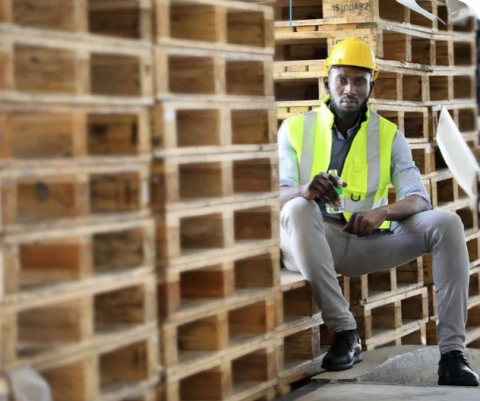 African American worker in safety vest suit and hardhat is drinking water during break while sitting on the pile of pallet wood at warehouse 